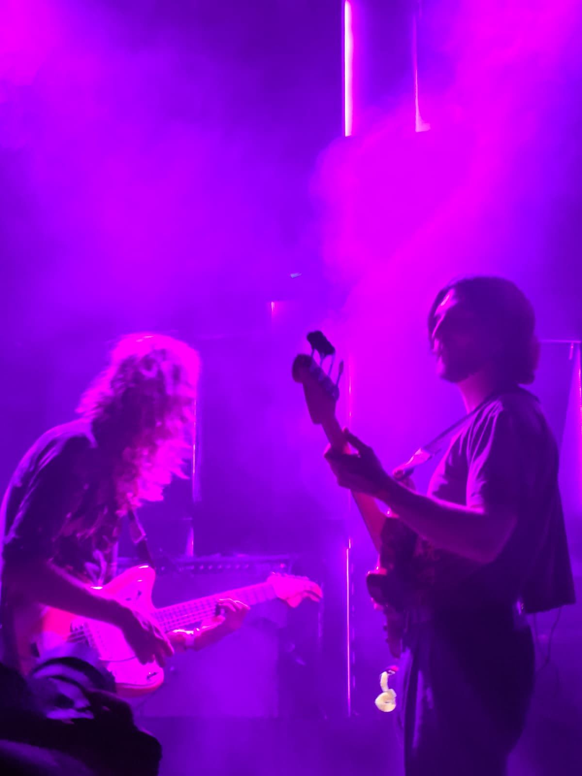 Two members of the YinYin band facing each other, playing guitar at a concert filled with fog under purple light